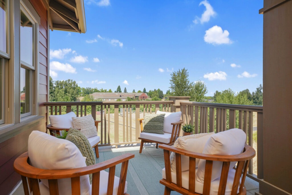 Alt text:
A furnished second-storey balcony with warm wood-framed chairs, white cushions and textured throw pillows overlooking a sunny tree-lined neighbourhood under a bright blue sky.