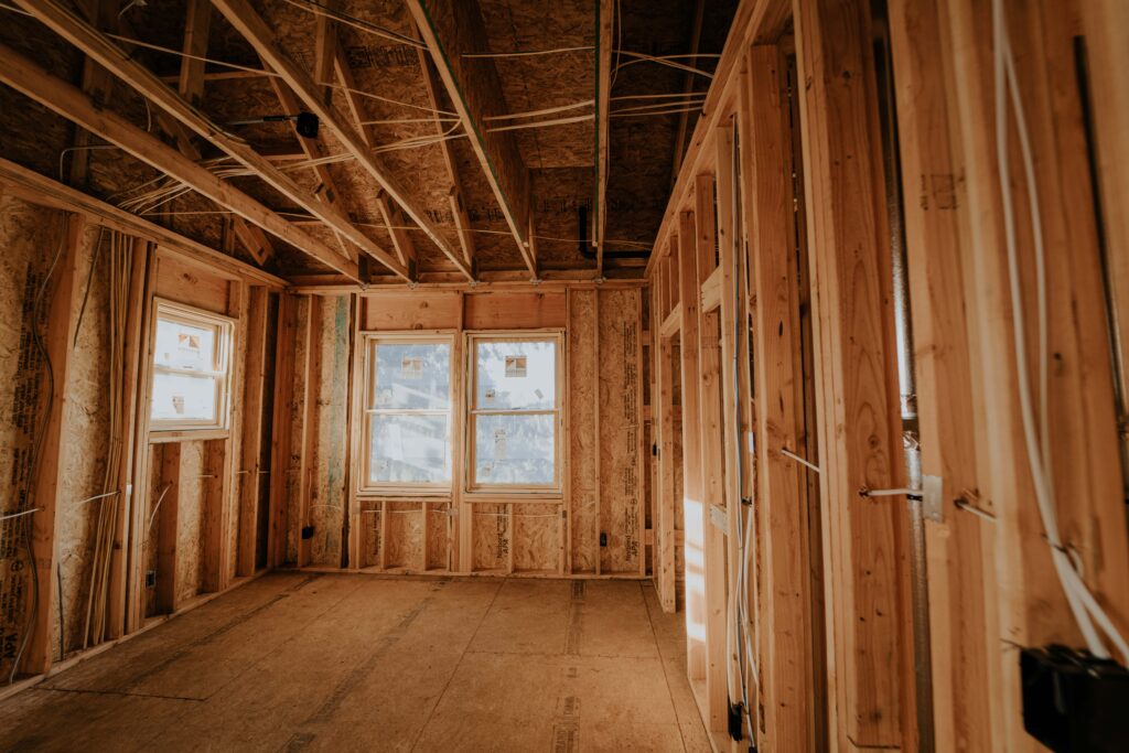 The interior framing of a new home under construction, with exposed wood studs, rough-in electrical wiring and subfloor visible throughout a sun-filled room.