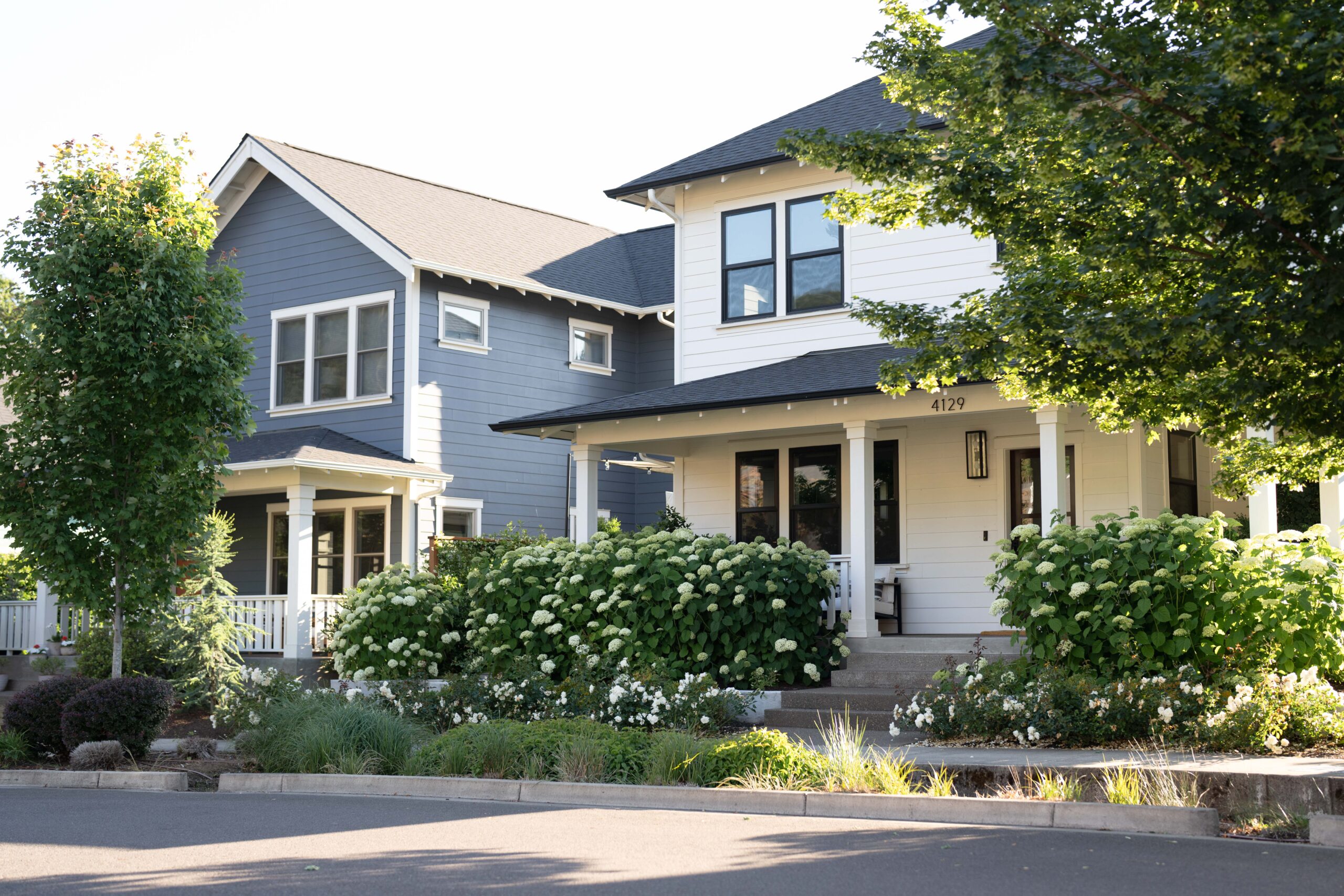 A charming two-storey craftsman-style home with slate blue and white exterior siding, black-framed windows and a covered front porch, surrounded by lush white hydrangea blooms and mature trees — finding the right lot starts with imagining a home like this in your neighbourhood.